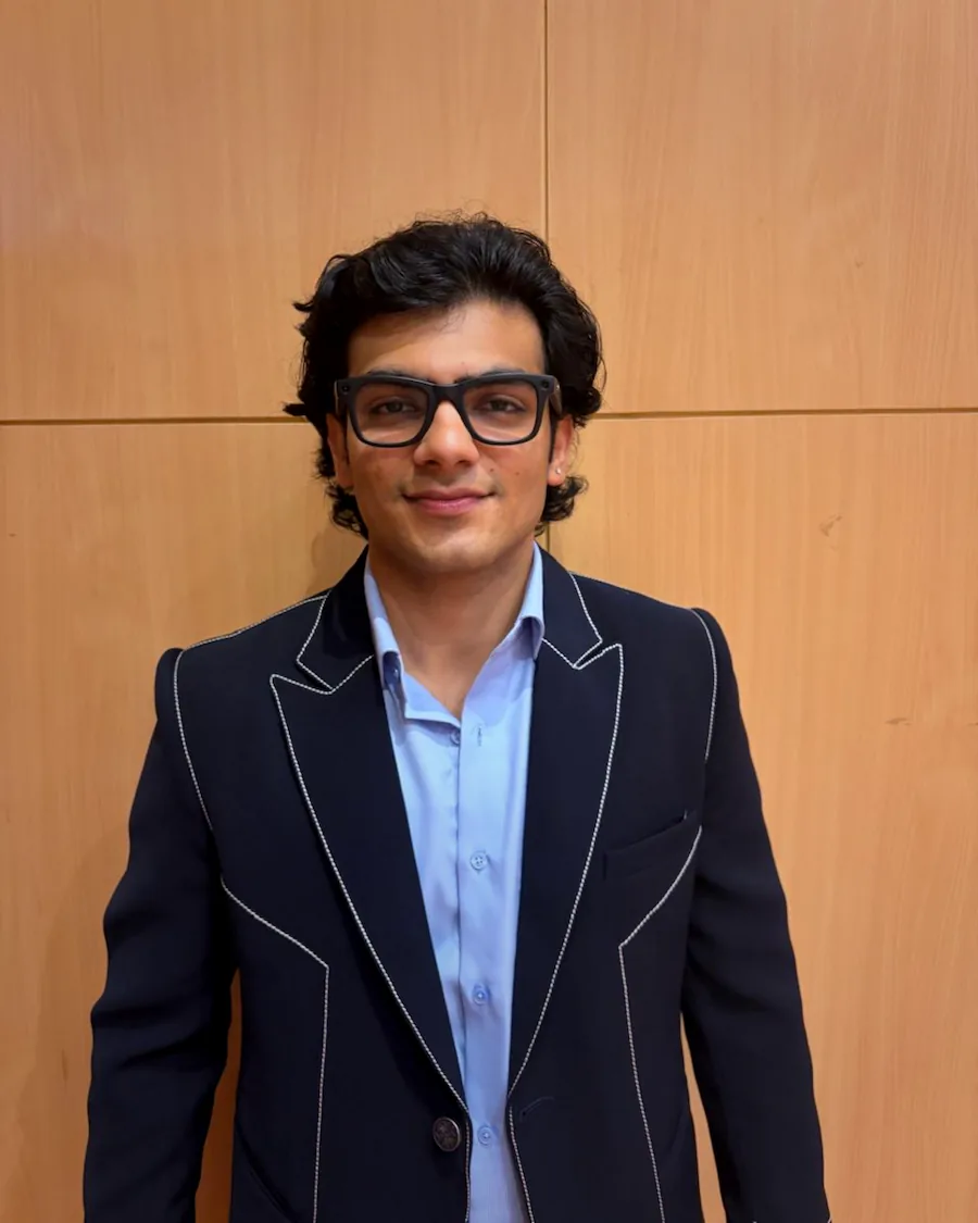 Saksham Goel, photographed against a pale wood-panelled wall in a navy jacket with white contrast stitching over a light blue shirt