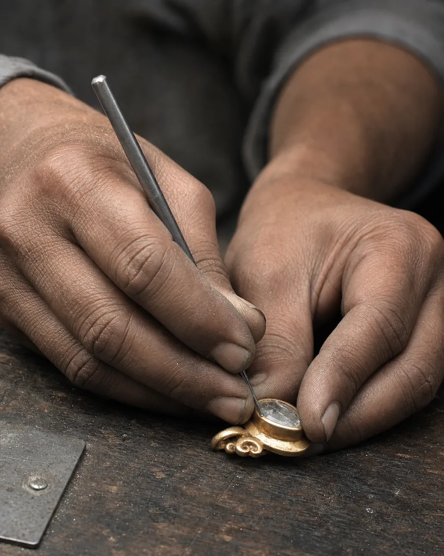 Two hands at a wooden bench, setting a rose-cut stone into a gold bezel with a steel tool